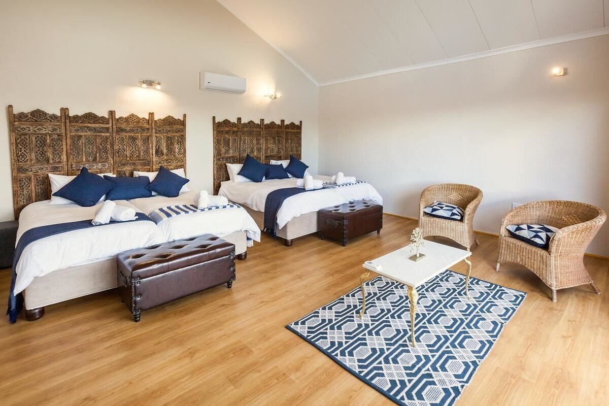 A hotel room with two beds, a bench, wicker chairs, a patterned rug, and a small table with a vase.