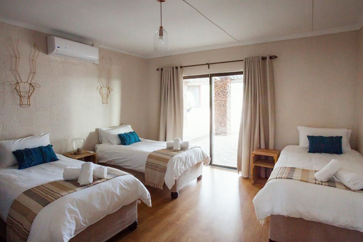 A hotel room with three beds, a large window with curtains, a wooden floor, and a hanging light fixture.