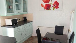 Een compacte keuken met blauwgroene kastjes, een magnetron, een waterkoker en een eethoek met een tafel en stoelen.