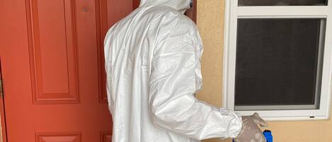 A person in a white protective suit using a blue device near a red door and a window.