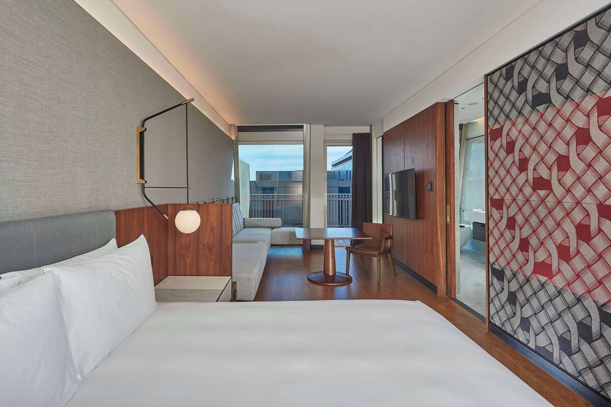 A modern hotel room with a large bed, a sitting area with a sofa and a table, and a view of the city through large windows.
