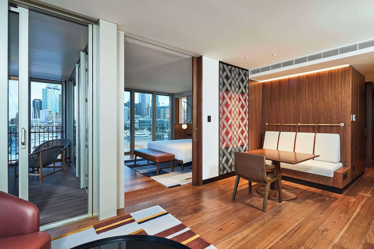 A modern hotel room with a large sliding glass door leading to a balcony, a sofa, a dining table, and a city view.
