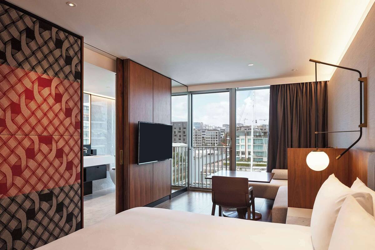 A modern hotel room with a large bed, a TV, a balcony with a view of the city, and a dining area.