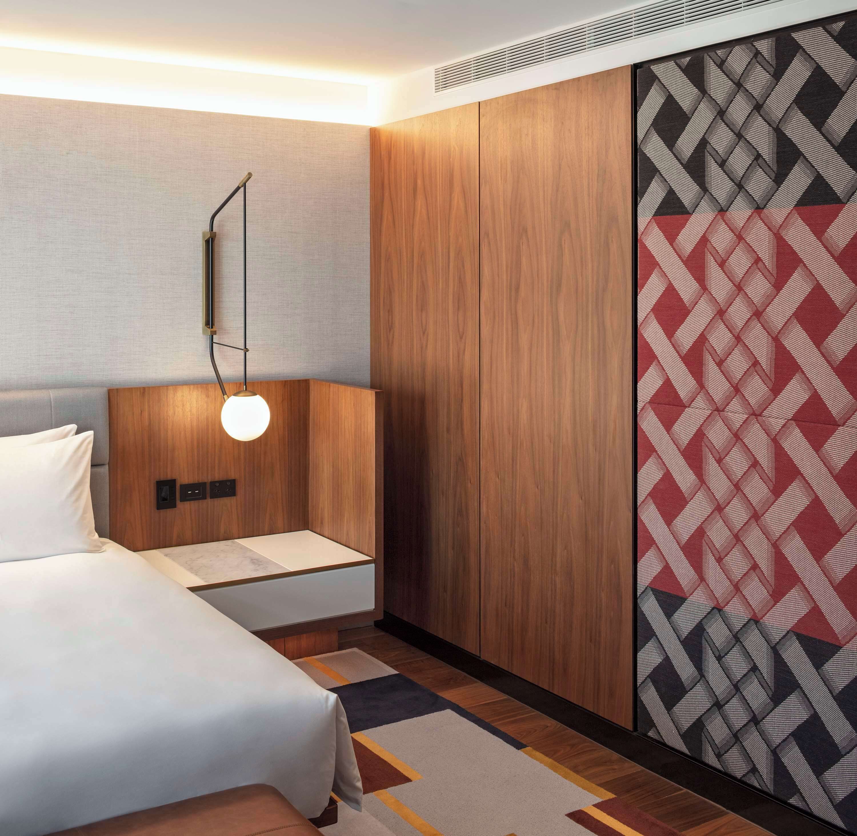 A modern hotel room with a bed, a bedside table, a wall-mounted lamp, and a patterned wall.
