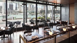 A restaurant with tables and chairs, large windows, and a view of a cityscape and water. - Park Hyatt Auckland (Auckland)