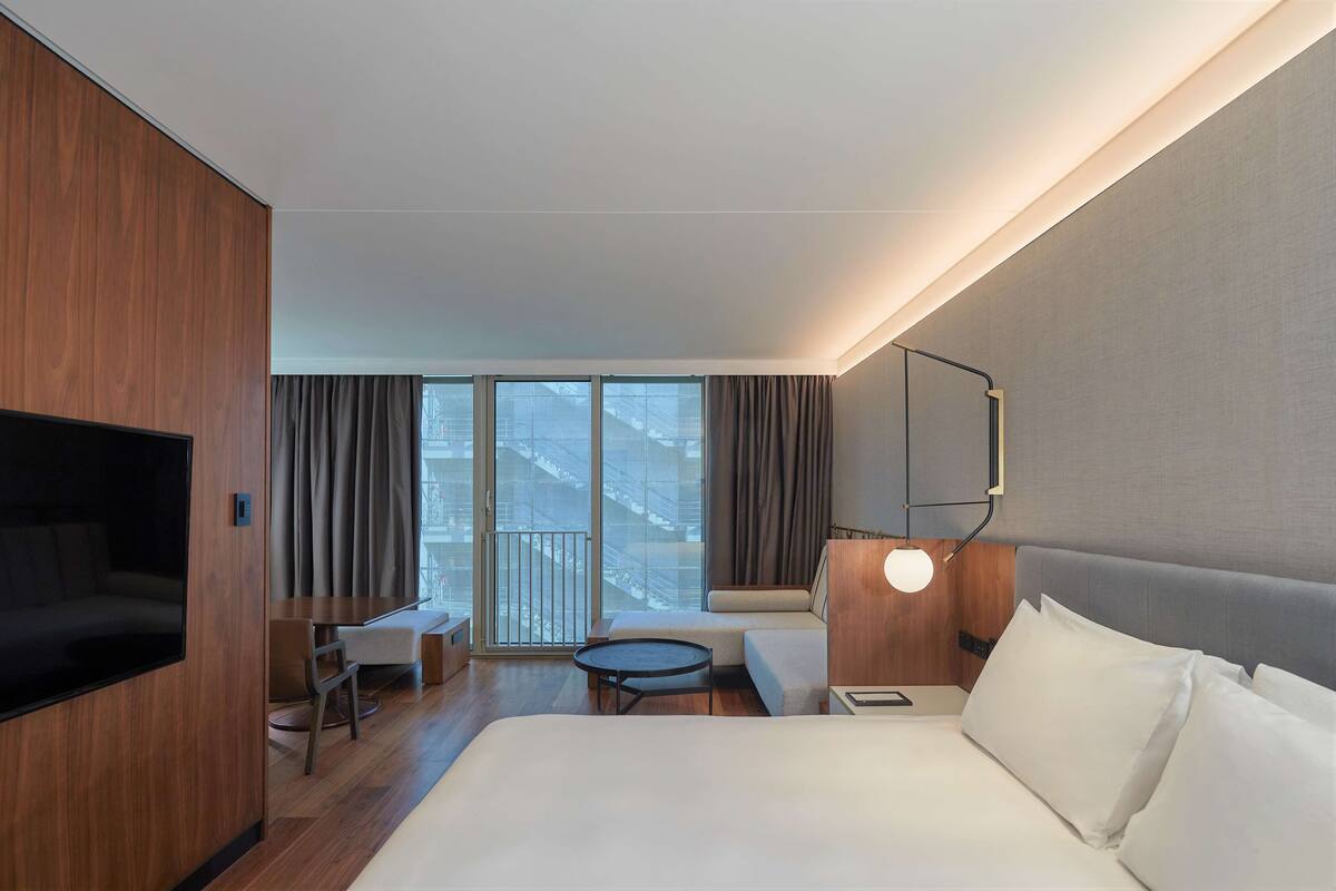 A modern hotel room with a large bed, a desk, a sofa, and a balcony with a view of the city.