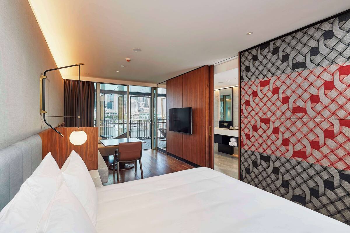A hotel room with a bed, a desk, a chair, a TV, and a large window offering a city view.