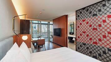A hotel room with a bed, a desk, a chair, a TV, and a large window offering a city view.