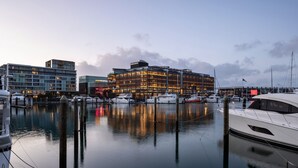 A marina with several yachts docked, a modern building with illuminated windows, and a calm water body reflecting the structures. - Park Hyatt Auckland (Auckland)
