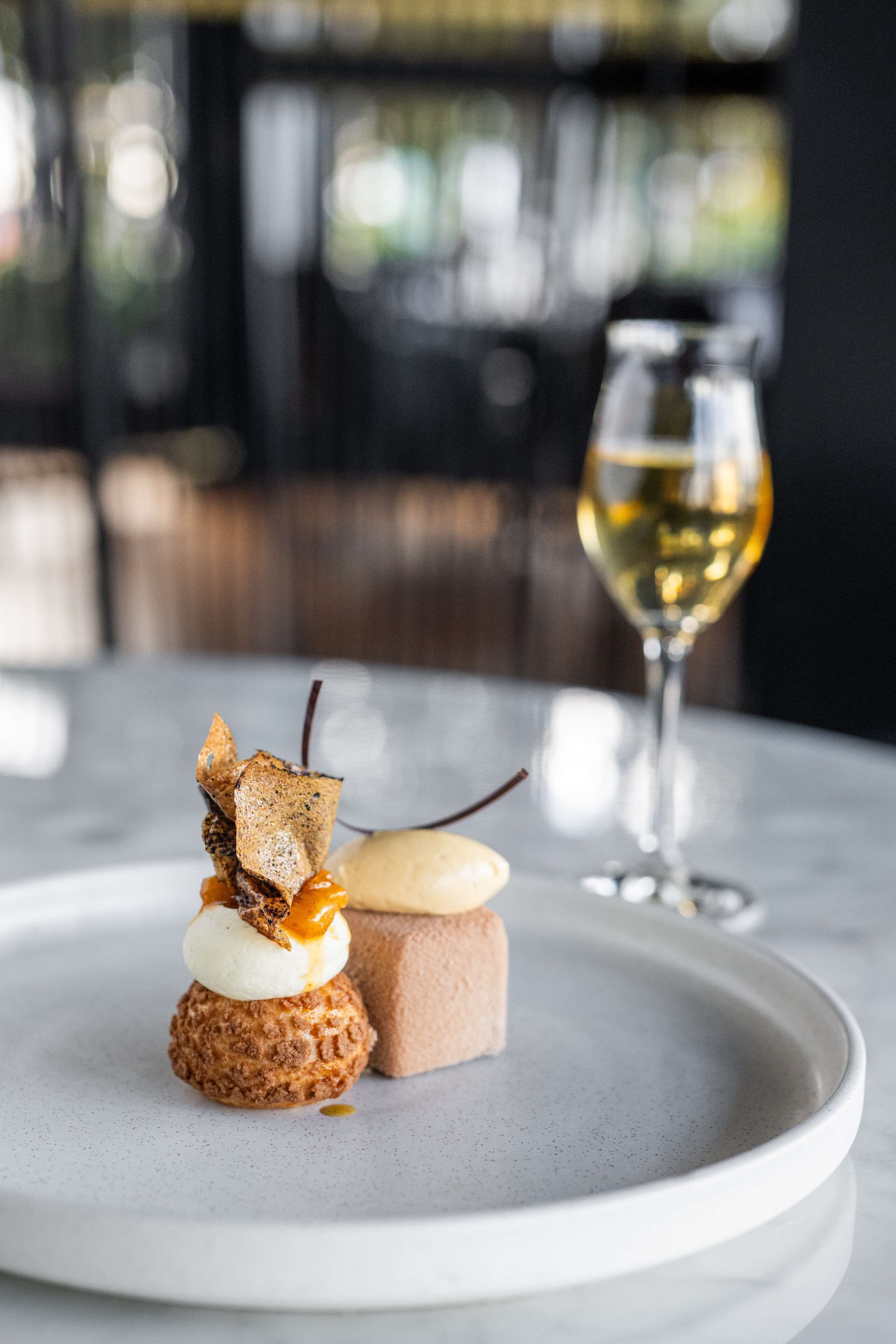 Ein Dessert auf einem weißen Teller, im Hintergrund ein Glas Weißwein.