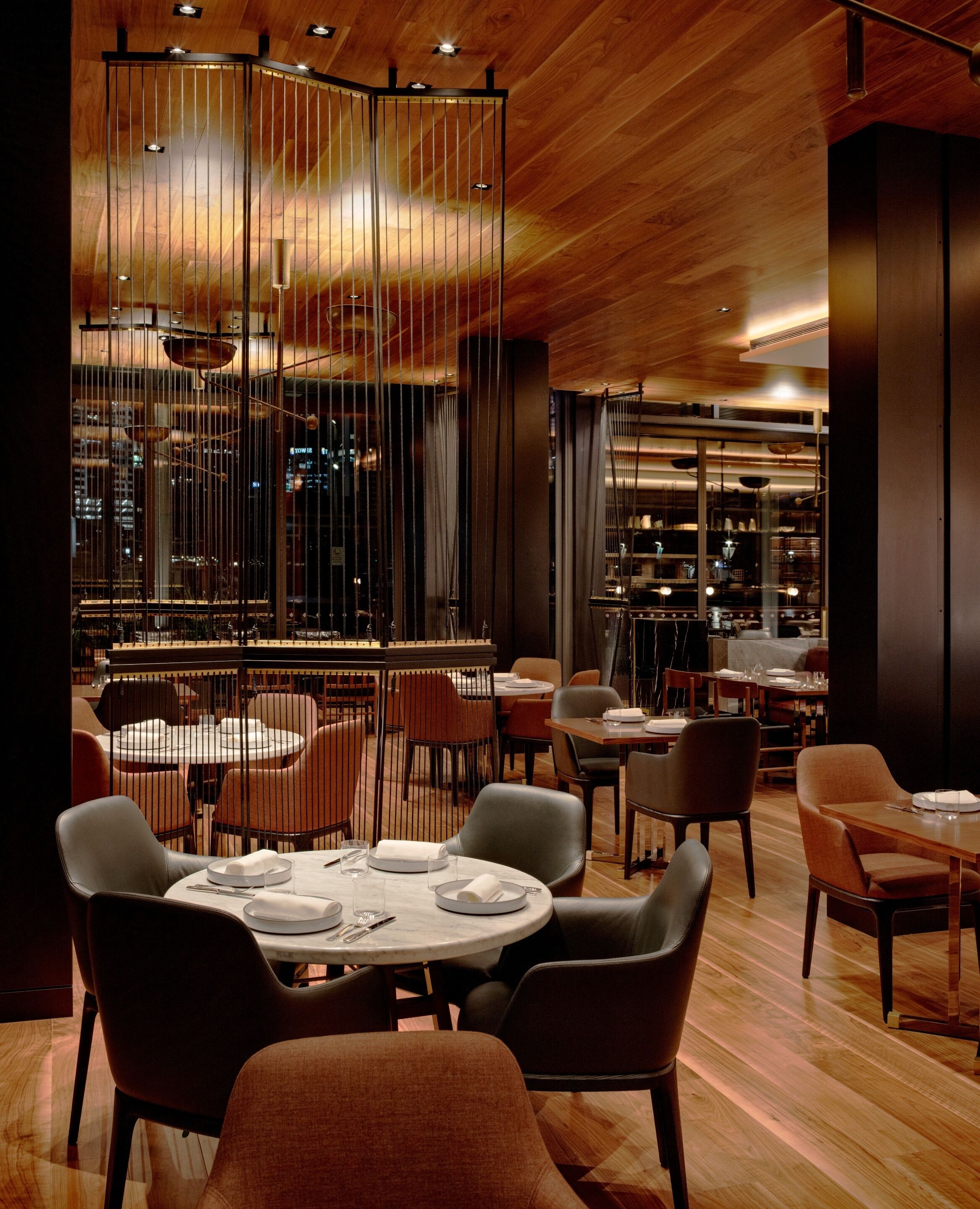 A modern restaurant with round tables and chairs, a wooden ceiling, and a city view through large windows.