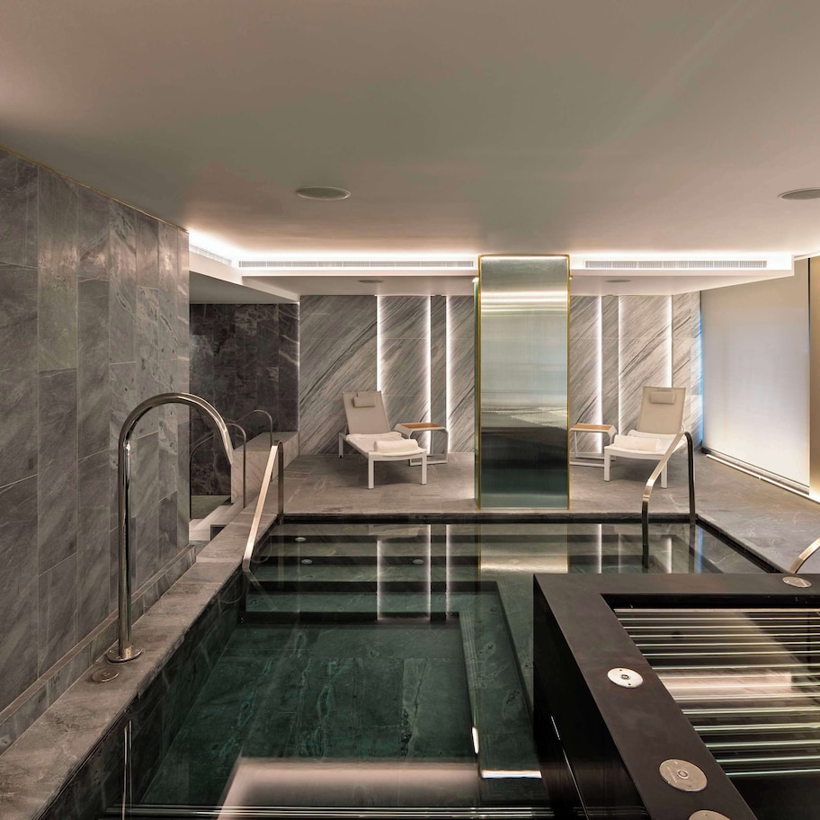 A modern indoor pool area with a glass-enclosed section, marble walls, and seating by the pool.