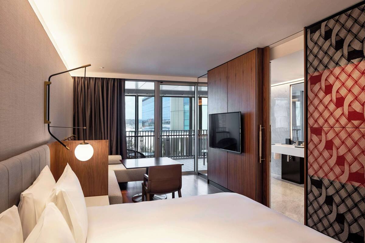 A hotel room with a bed, a desk, a chair, a TV, and a balcony with a view of buildings.