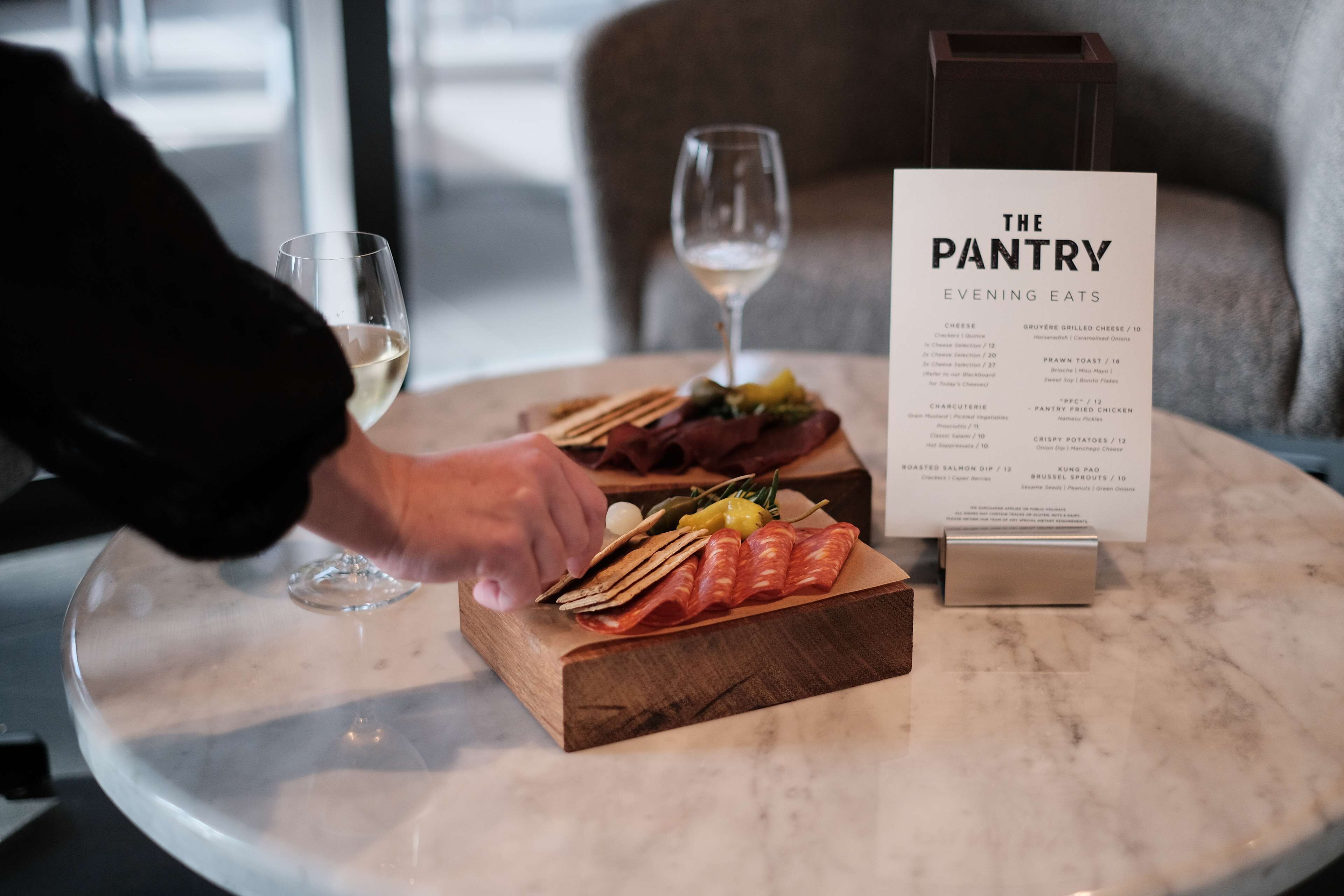 A menu titled "The Pantry Evening Eats" on a table with a glass of white wine and a wooden board with various food items.