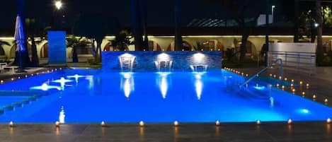 Uma piscina noturna com fontes iluminadas e um deck revestido com azulejos.
