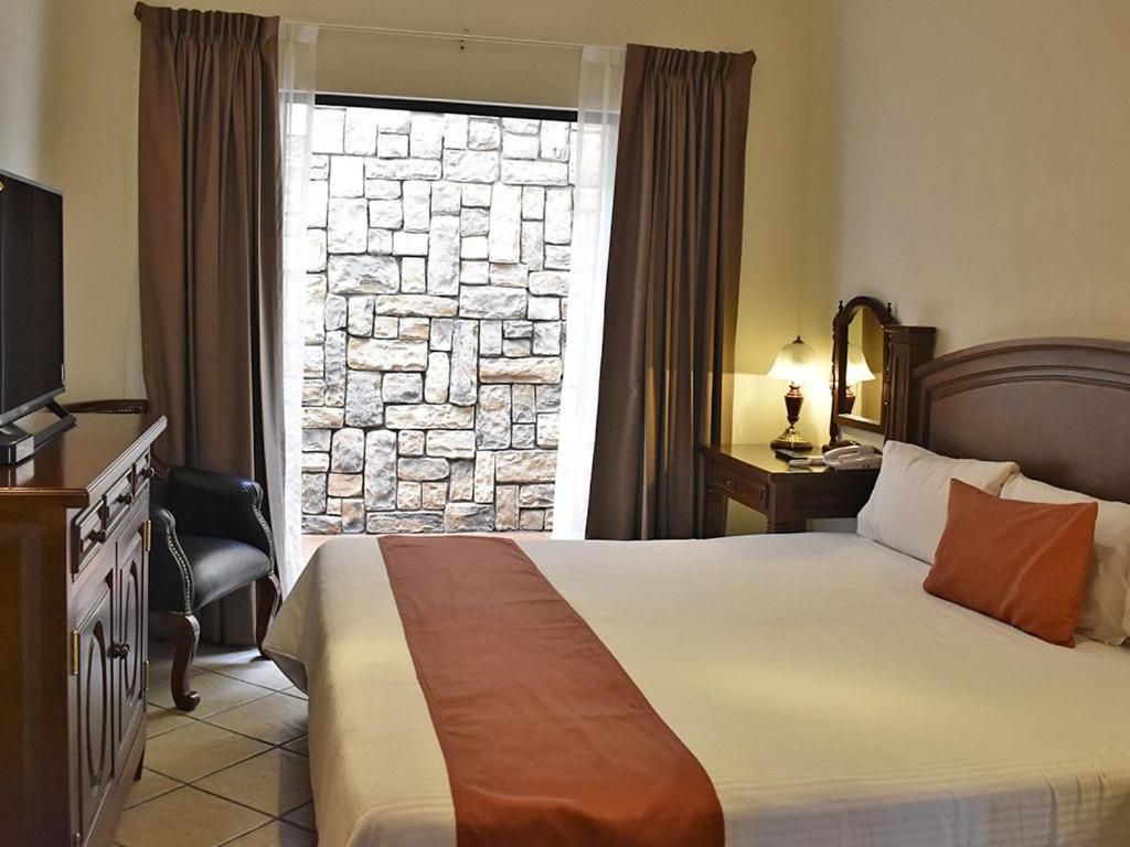 Una habitación de hotel con cama, escritorio, silla, mesita de noche con lámpara y pared de piedra.