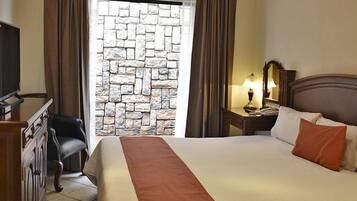 Una habitación de hotel con cama, escritorio, silla, mesita de noche con lámpara y pared de piedra.