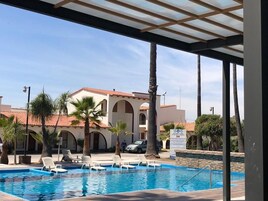Una piscina con sillas de descanso, palmeras y un edificio con ventanas arqueadas al fondo.