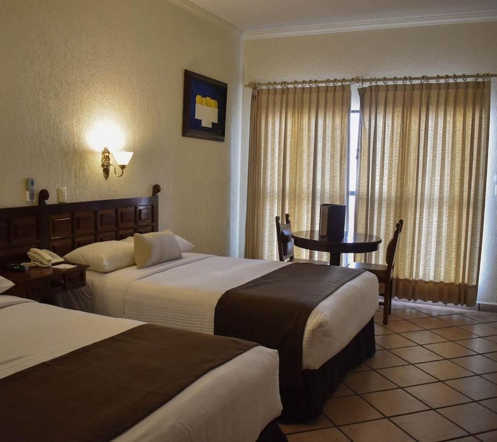 A hotel room with two beds, a small table, a chair, a lamp, and a window with curtains.