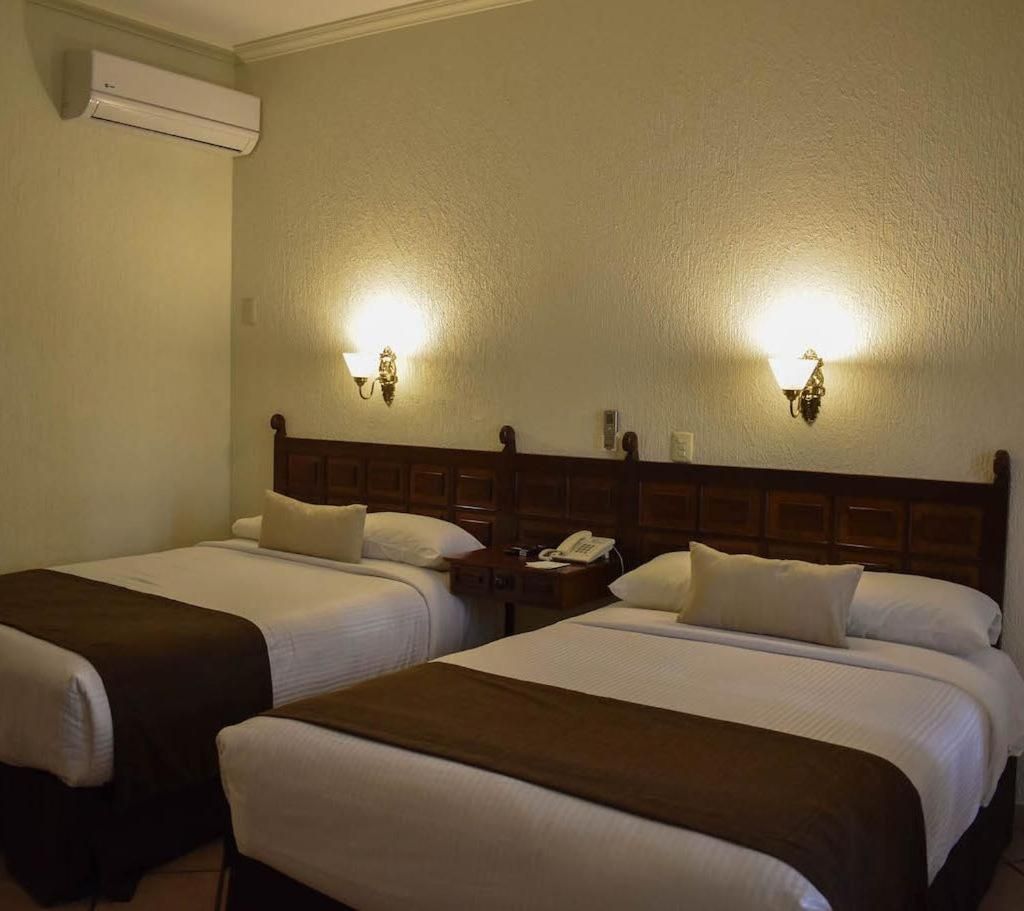 A hotel room with two beds, each with a wooden headboard and white bedding. There are wall-mounted light fixtures above each bed.