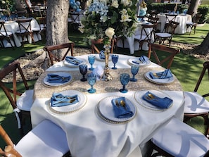Un área de comedor al aire libre con mesas redondas dispuestas para una comida, con manteles blancos, servilletas y copas azules.