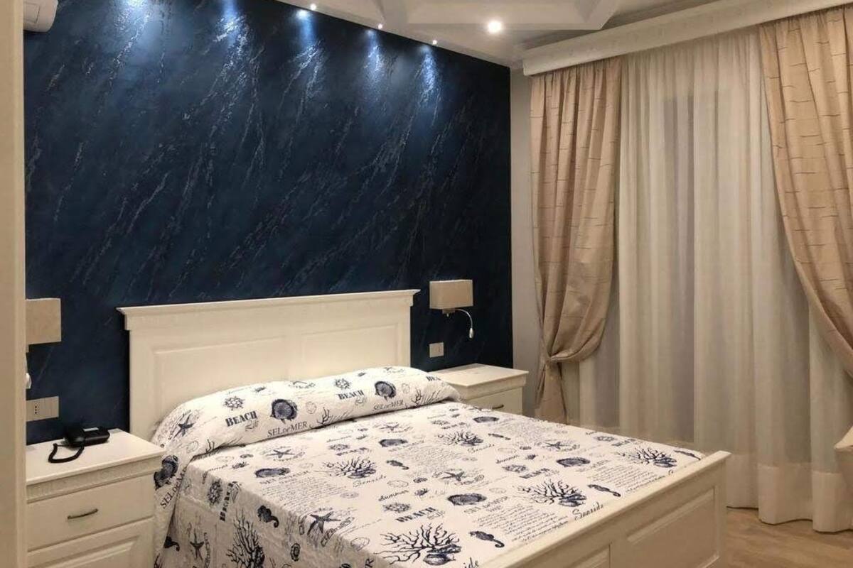 Ein Schlafzimmer mit dunkelblauer Marmorwand, einem weißen Bett mit gemustertem Überwurf, einem Nachttisch und einem Fenster mit beigen Vorhängen.