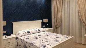 A bedroom with a dark blue marble wall, a white bed with a patterned bedspread, a bedside table, and a window with beige curtains. - Stella del Mare (Licata)