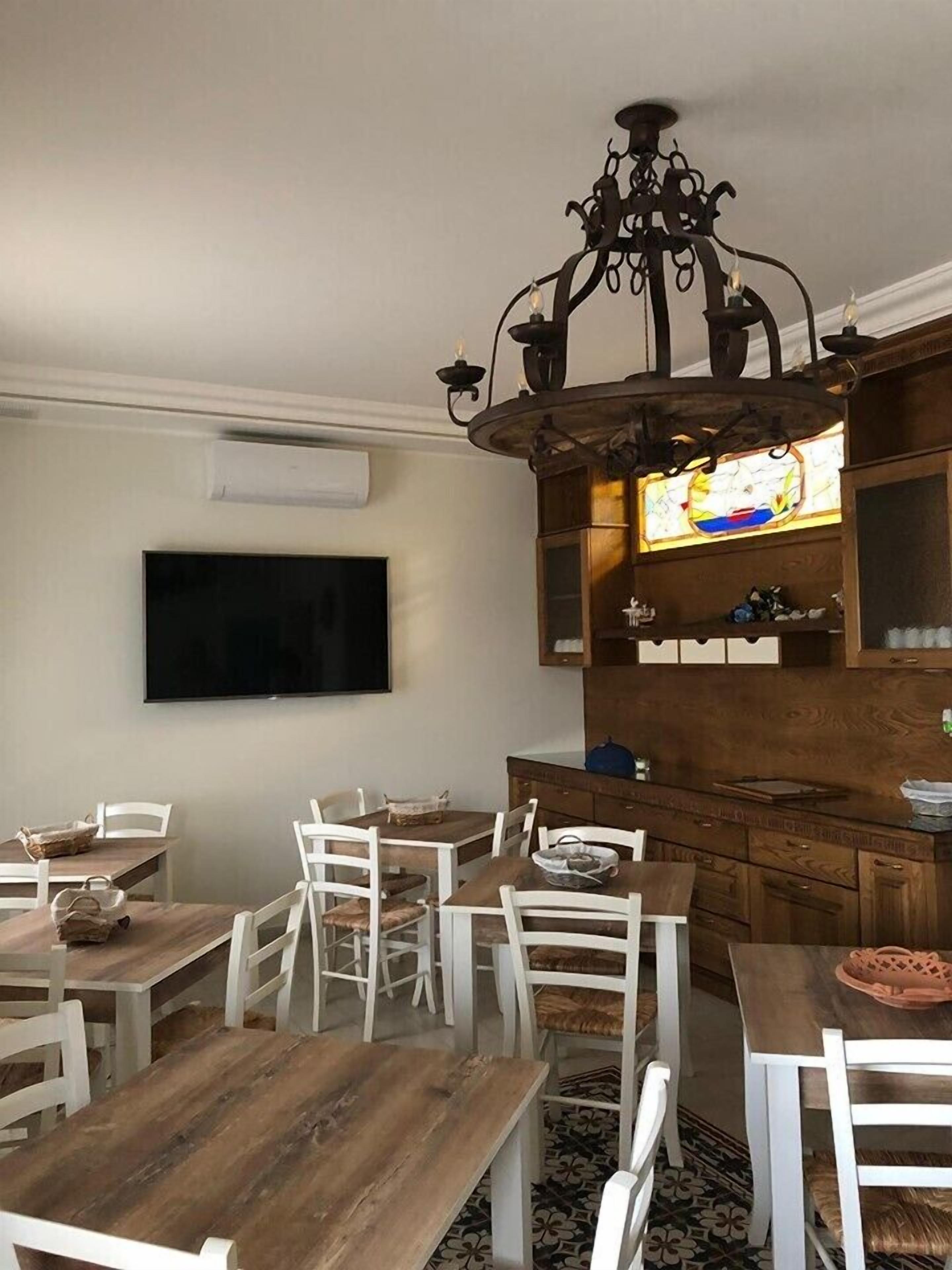A dining area with wooden tables and white chairs, a chandelier, a flat-screen TV, and a wooden cabinet with shelves.