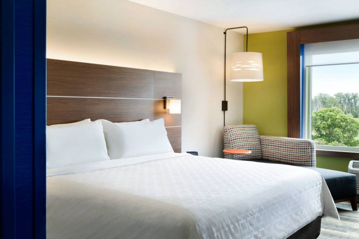 A hotel room with a large bed, a chair, a lamp, and a window with a view of trees.