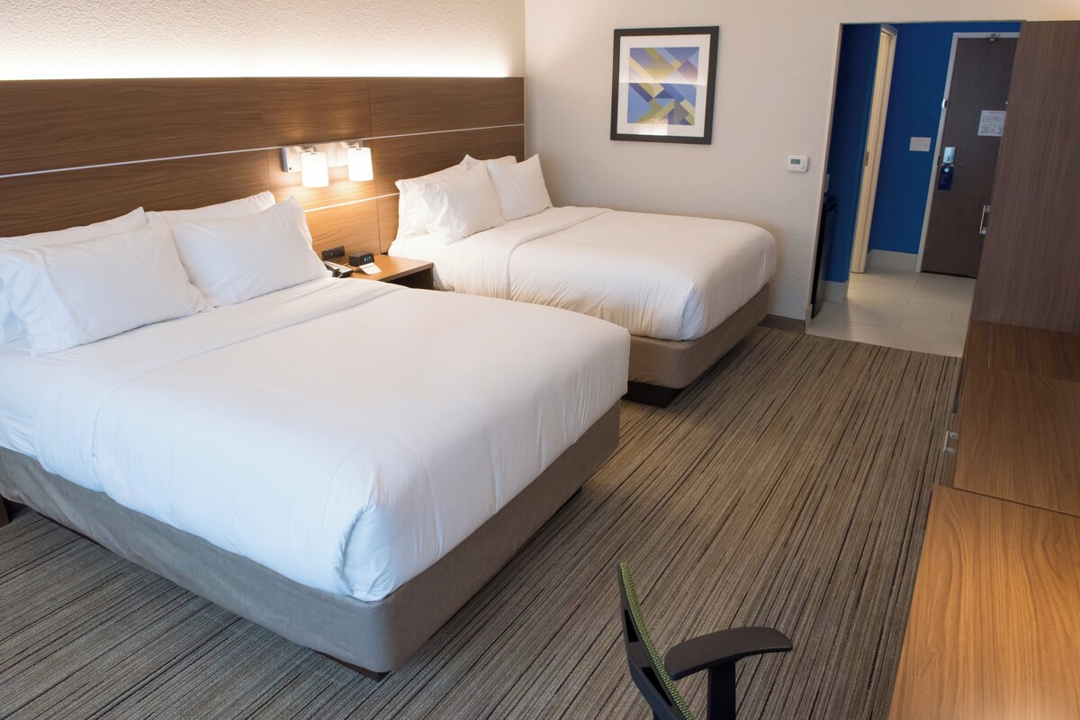 A hotel room with two beds, a nightstand, a lamp, a framed picture on the wall, and a door leading to another room.