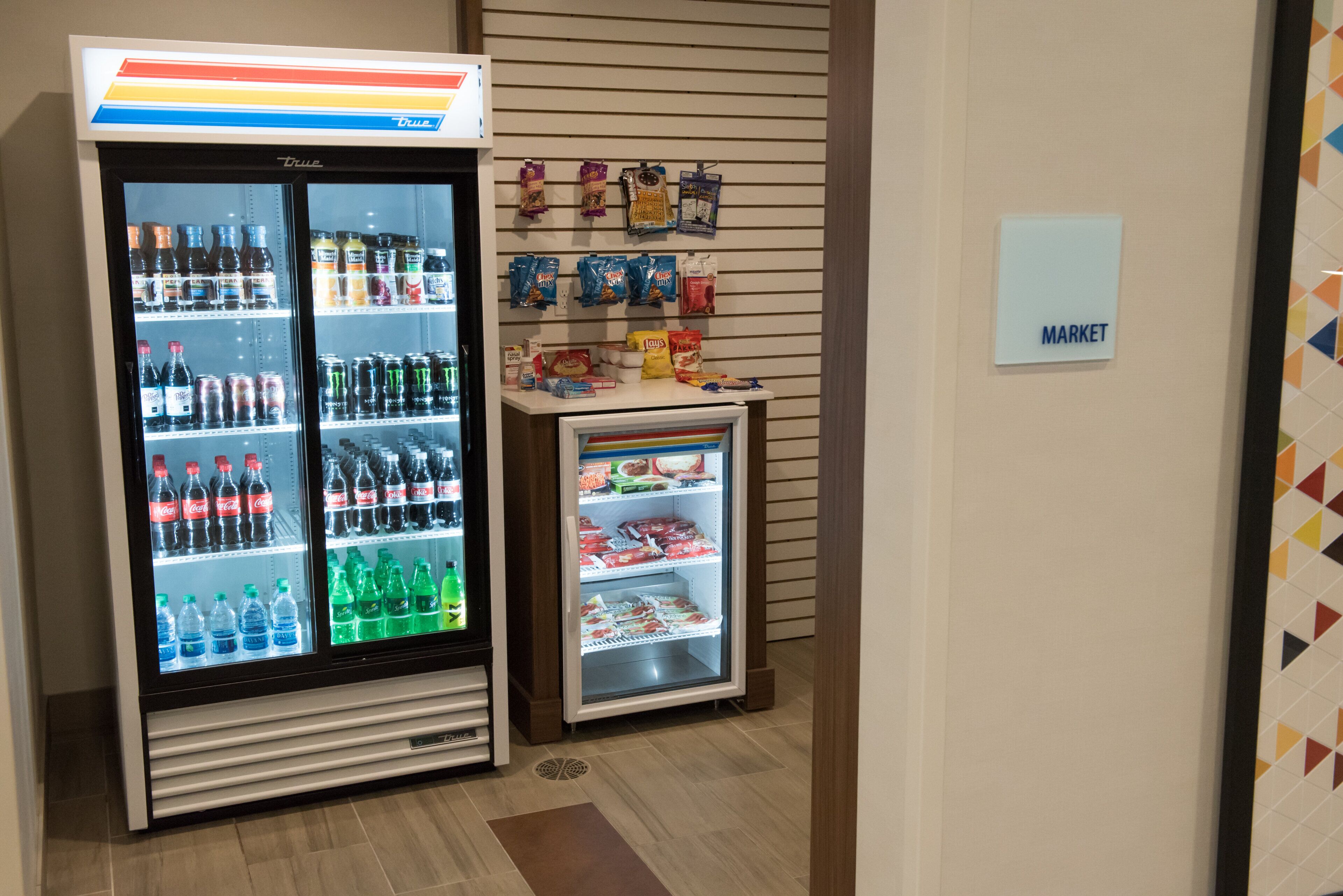 Una máquina expendedora con diferentes bebidas y aperitivos, un refrigerador pequeño con productos congelados, y un cartel que indica un área de mercado.