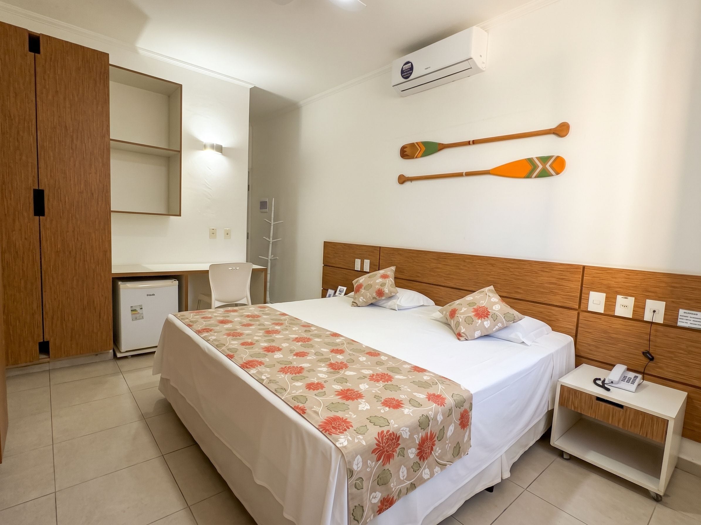 A hotel room with a bed, a desk, a refrigerator, a wall-mounted air conditioner, and two decorative paddles on the wall.