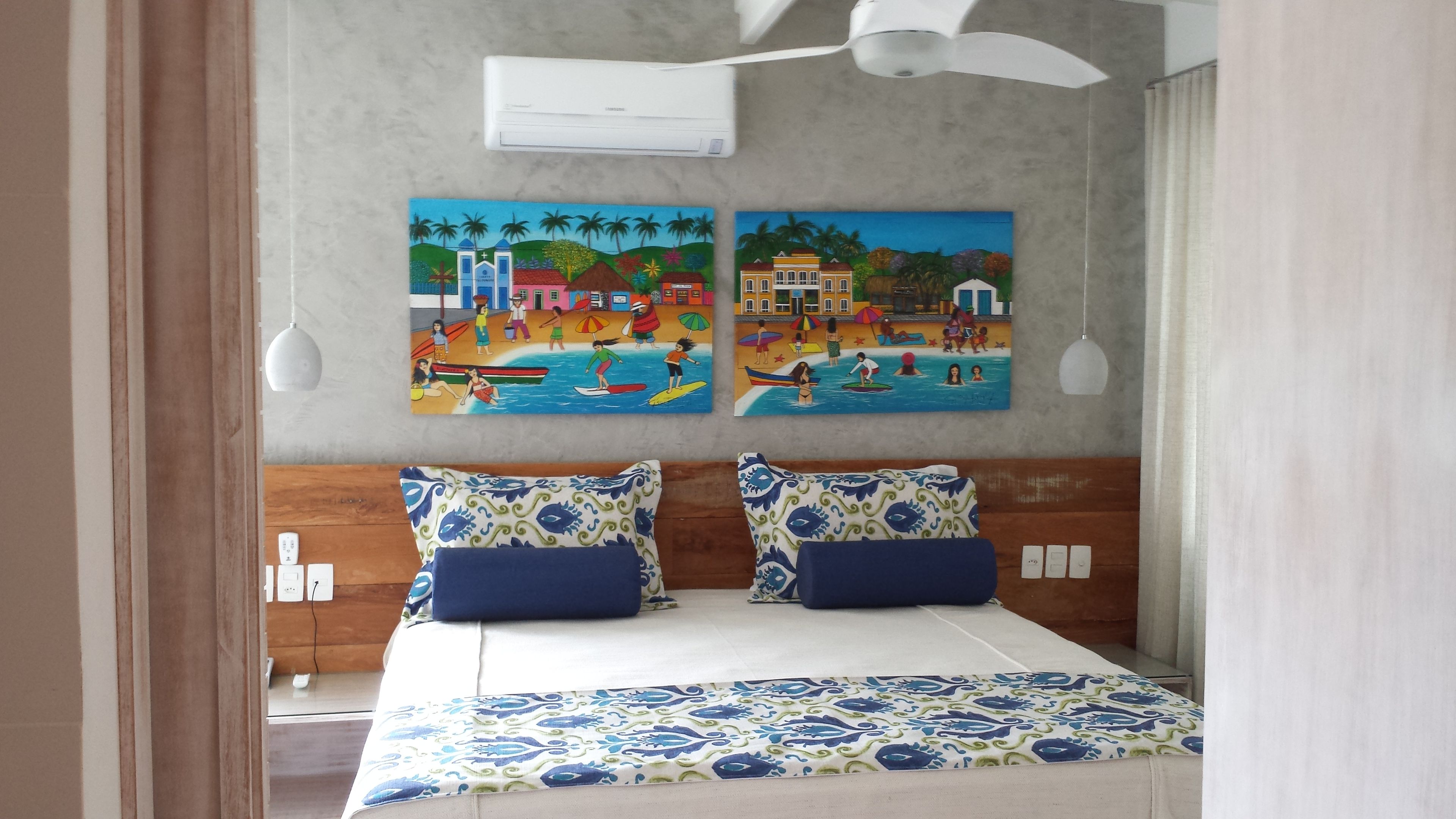 A bedroom with a bed, two wall-mounted air conditioners, and two colorful paintings depicting beach scenes.