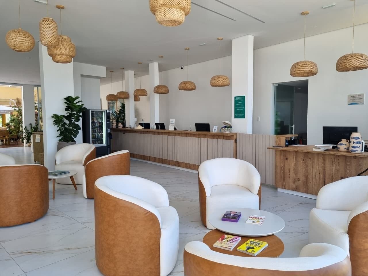 Un área de recepción moderna con sillones tapizados en blanco y marrón, un mostrador de recepción de madera y lámparas colgantes tejidas.