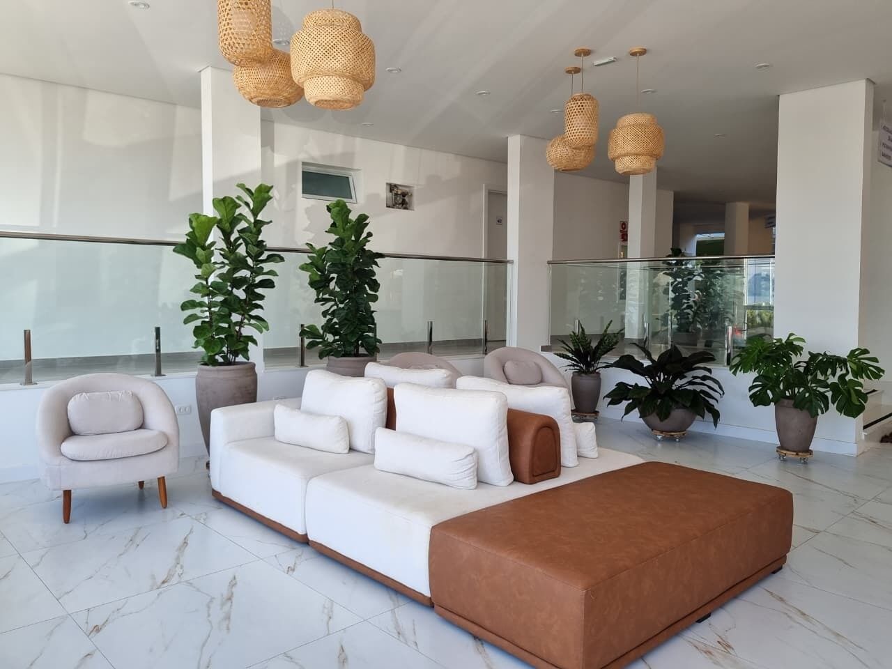 A modern living room with a white sofa, a brown ottoman, and a white armchair. There are several potted plants and wicker pendant lights.