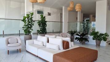 A modern living room with a white sofa, a brown ottoman, and a white armchair. There are several potted plants and wicker pendant lights.