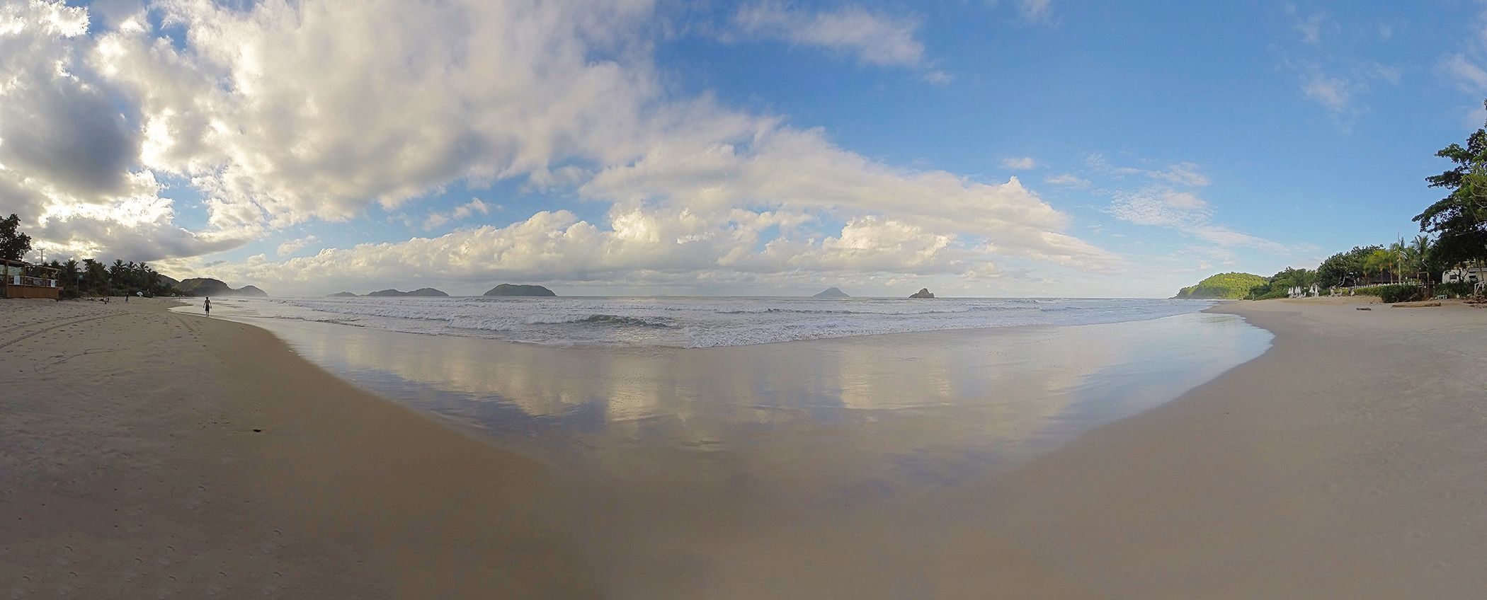 A beach with a wide sandy shore, calm waves, and a clear sky with scattered clouds.