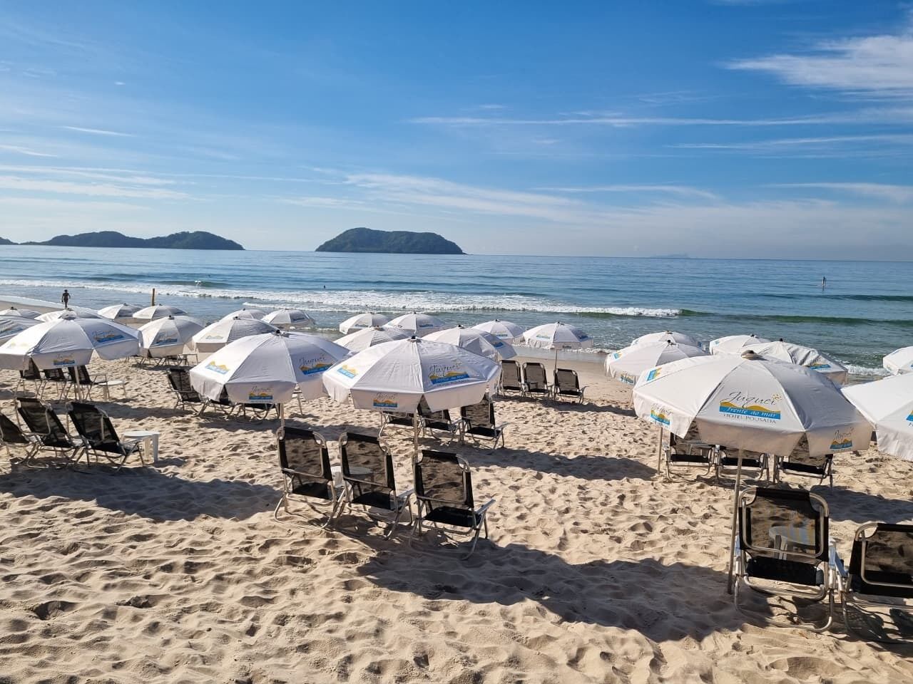Une plage bordée de rangées de chaises longues et de parasols, un rivage sablonneux et une mer calme avec des îles à l’horizon.