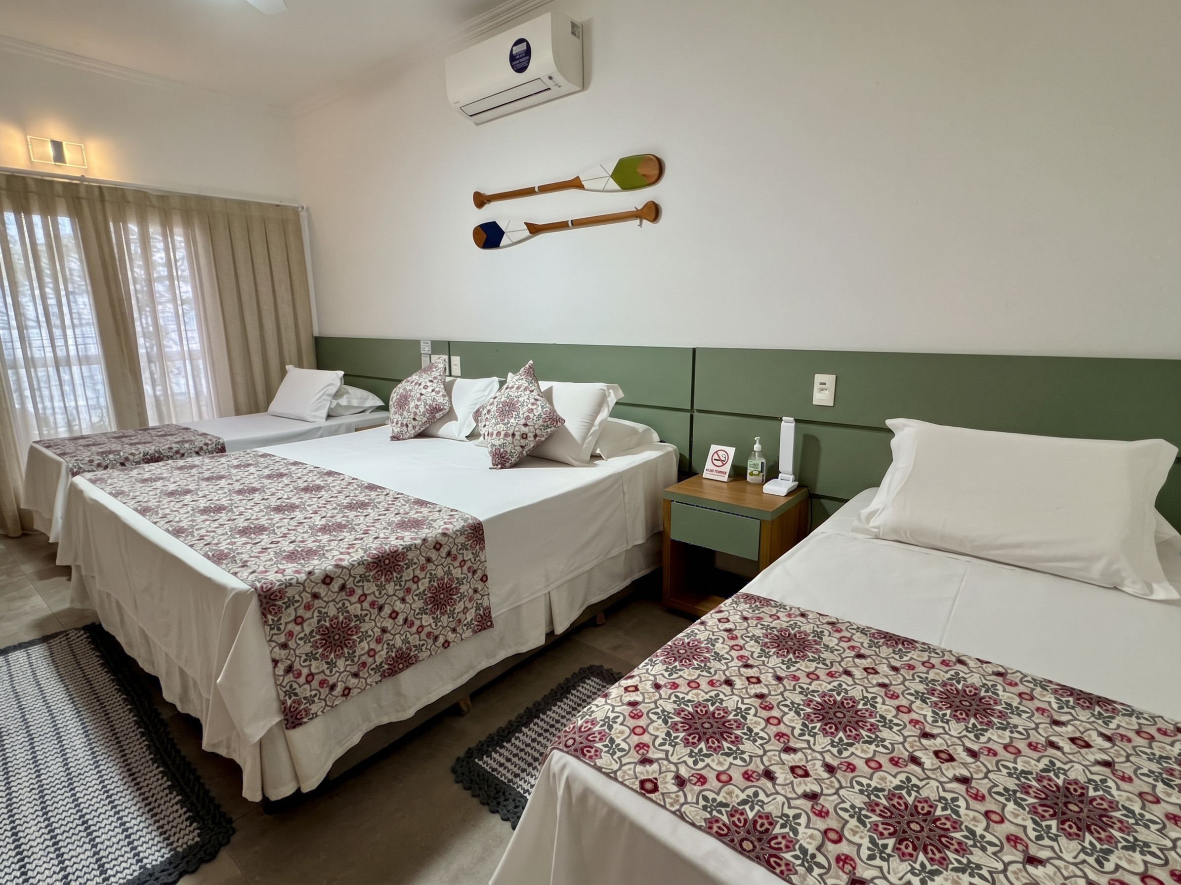 A hotel room with three beds, a nightstand, a wall-mounted air conditioner, and a window with curtains.