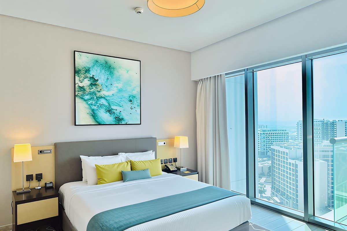 Ein modernes Hotelzimmer mit einem großen Bett, zwei Nachttischen mit Lampen, Blick auf die Stadt durch raumhohe Fenster und einem großen abstrakten Gemälde an der Wand.