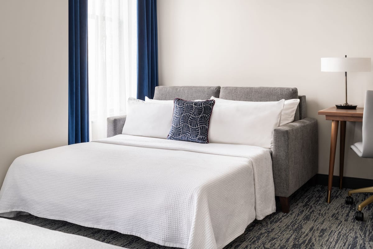 A hotel room with a large bed, a grey armchair, a small table with a lamp, and a window with blue curtains.