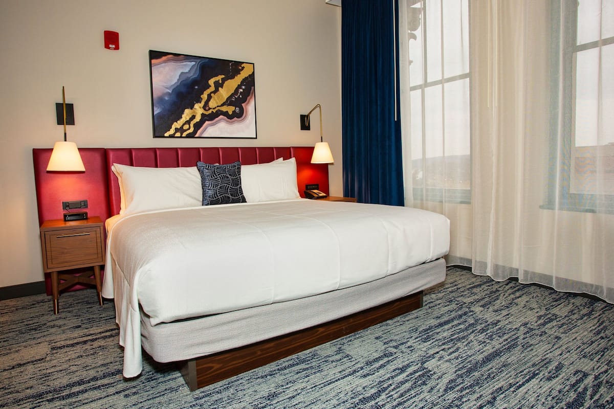 A hotel room with a large bed, two bedside tables with lamps, a red headboard, a framed abstract painting, and a window with blue curtains.