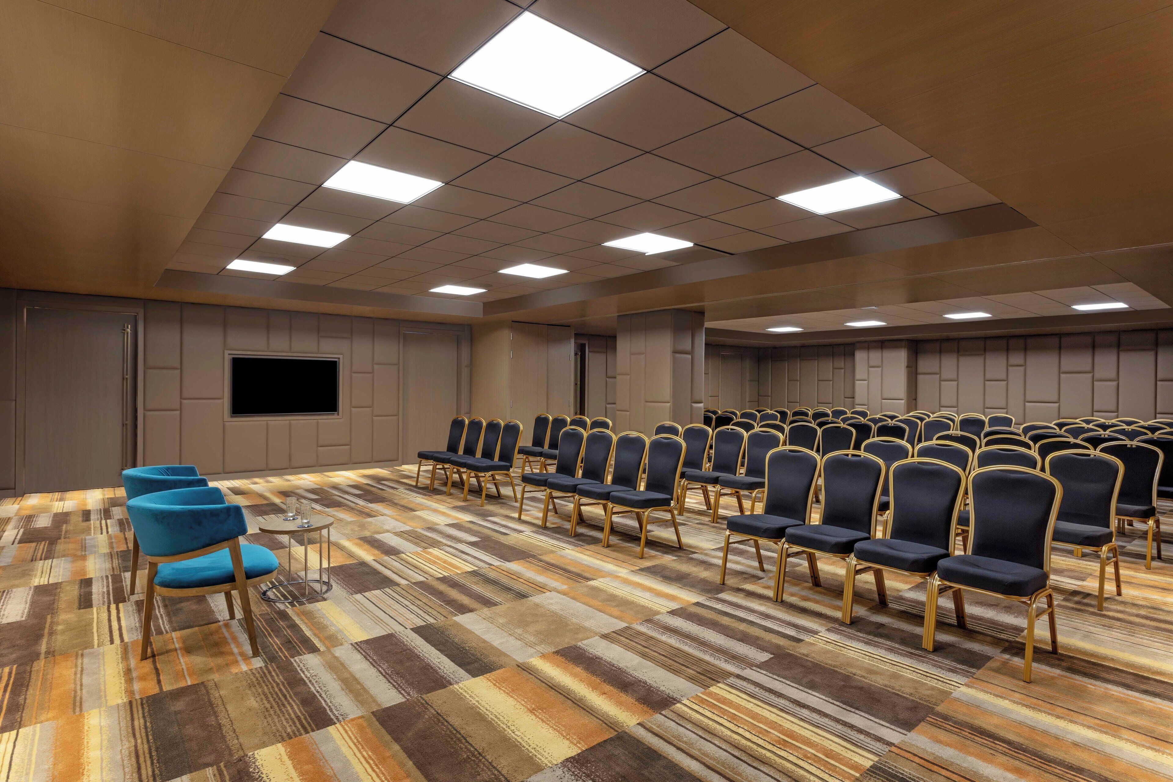 Un salón de conferencias con filas de sillas, piso alfombrado y una televisión de pantalla plana en la pared.