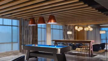 Un espace moderne dédié aux tables de billard, situé à l’intérieur, avec une table de billard entourée de chaises et de tables, et une grande fenêtre qui laisse entrer la lumière naturelle.