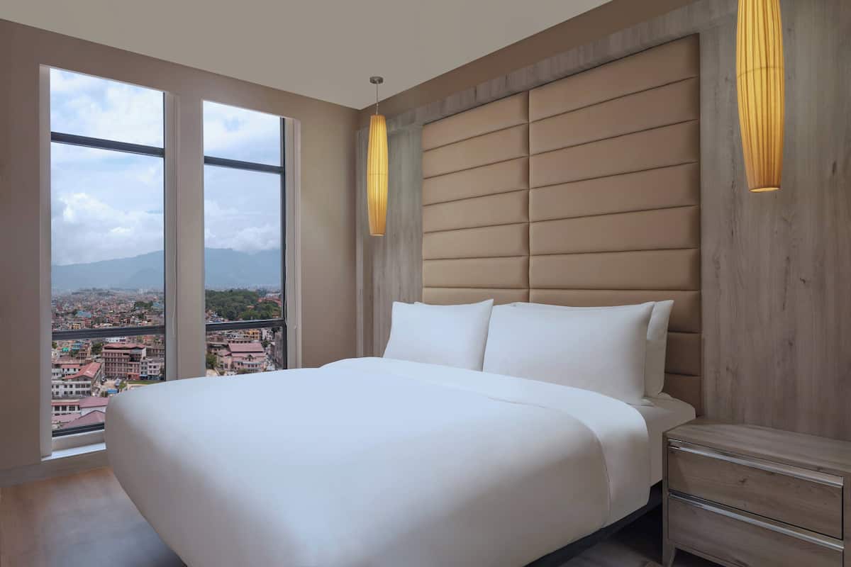 Une chambre avec un grand lit, une tête de lit en bois, une table de chevet et une vue sur le paysage urbain, visible à travers de grandes fenêtres.