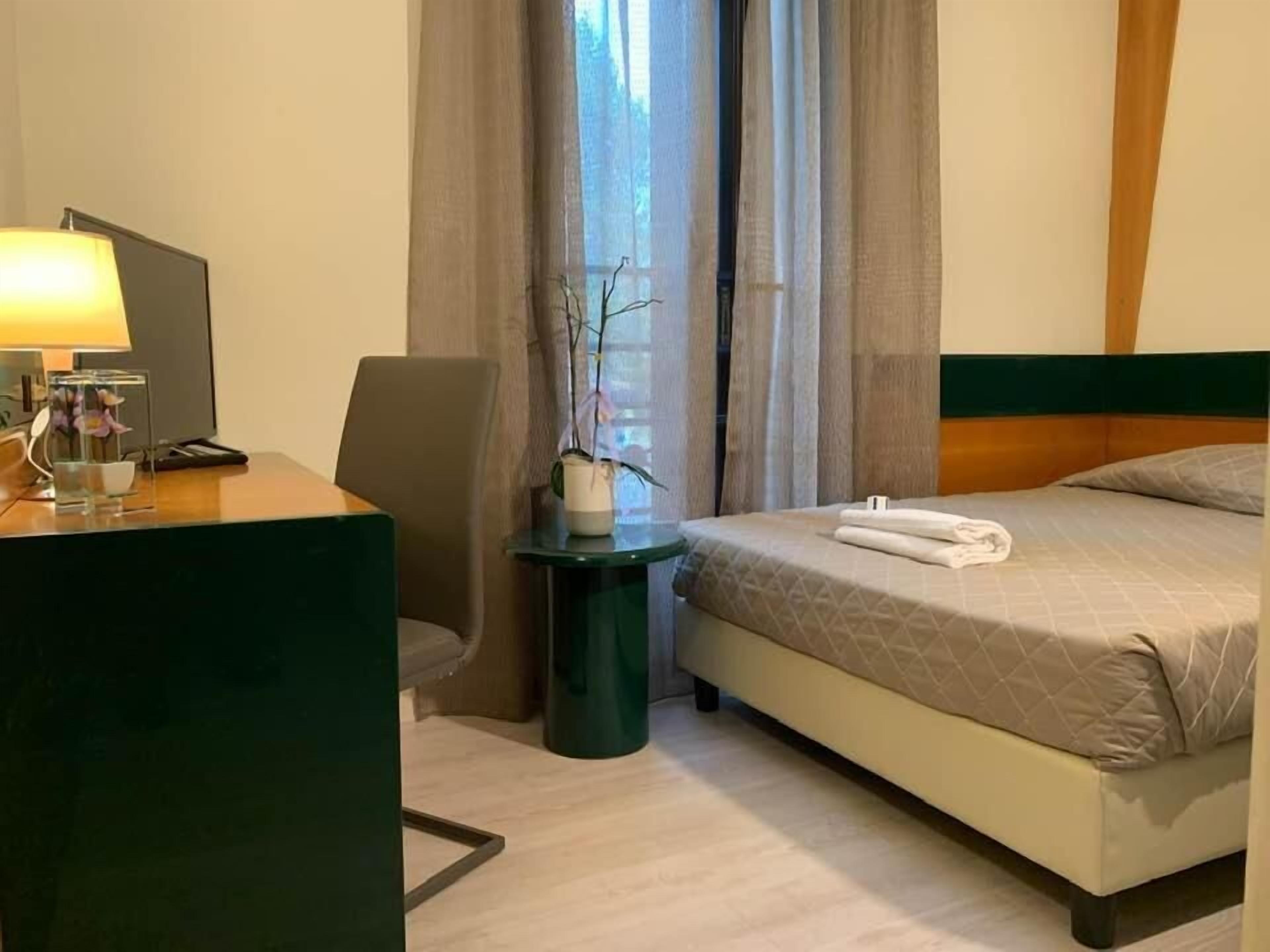 A hotel room with a bed, a desk with a lamp, a chair, and a small table with a vase.