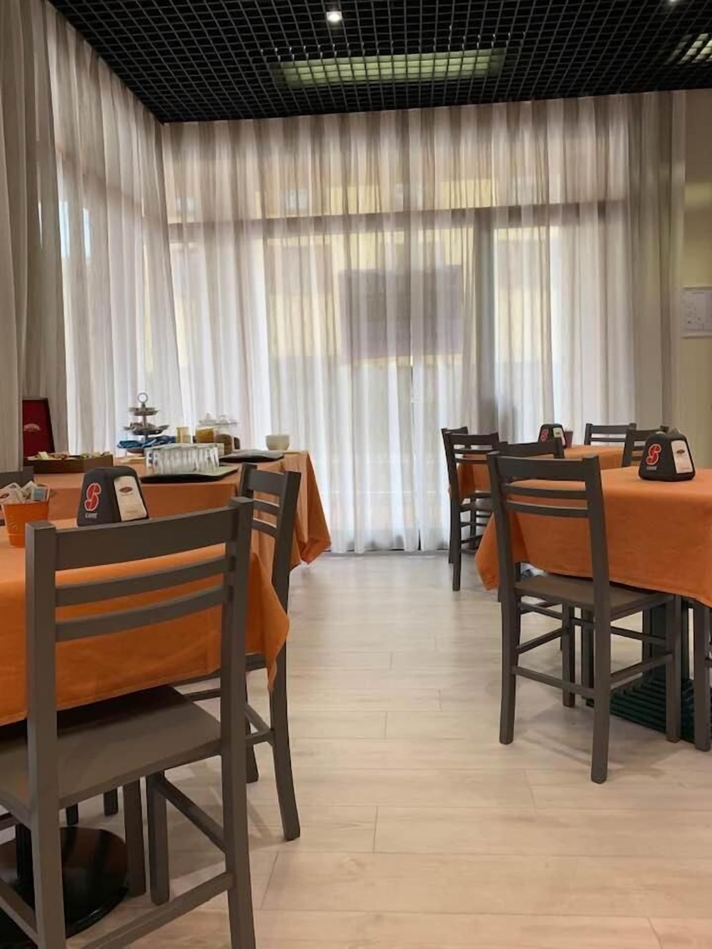 A dining area with tables covered in orange tablecloths, wooden chairs, and a backdrop of sheer white curtains.