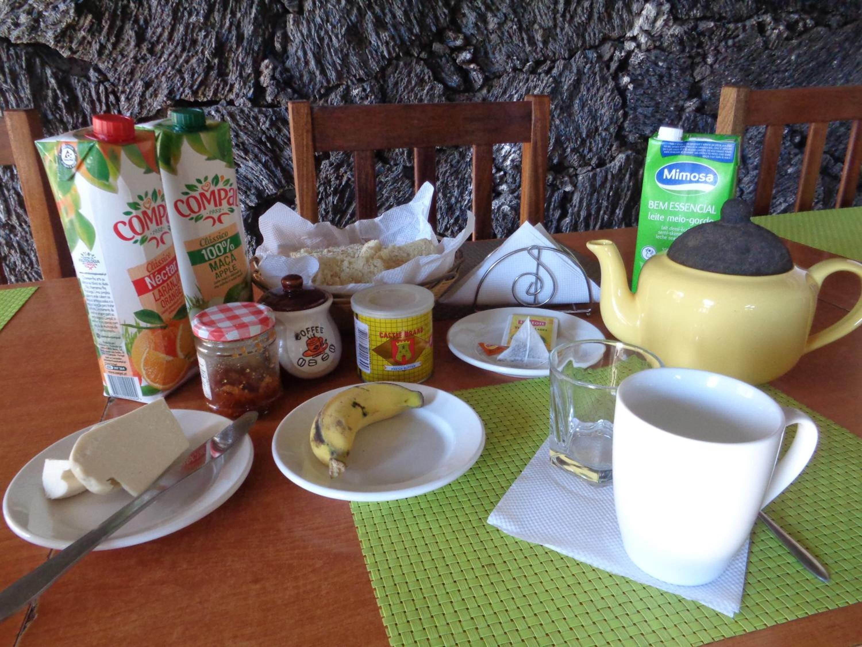 Una mesa de desayuno con diferentes alimentos y bebidas, incluyendo un cartón de jugo de naranja, una botella de leche, un frasco de mermelada, un plátano, un plato con mantequilla, una taza de té y una tetera.