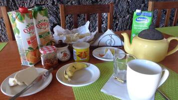 Una mesa de desayuno con diferentes alimentos y bebidas, incluyendo un cartón de jugo de naranja, una botella de leche, un frasco de mermelada, un plátano, un plato con mantequilla, una taza de té y una tetera.