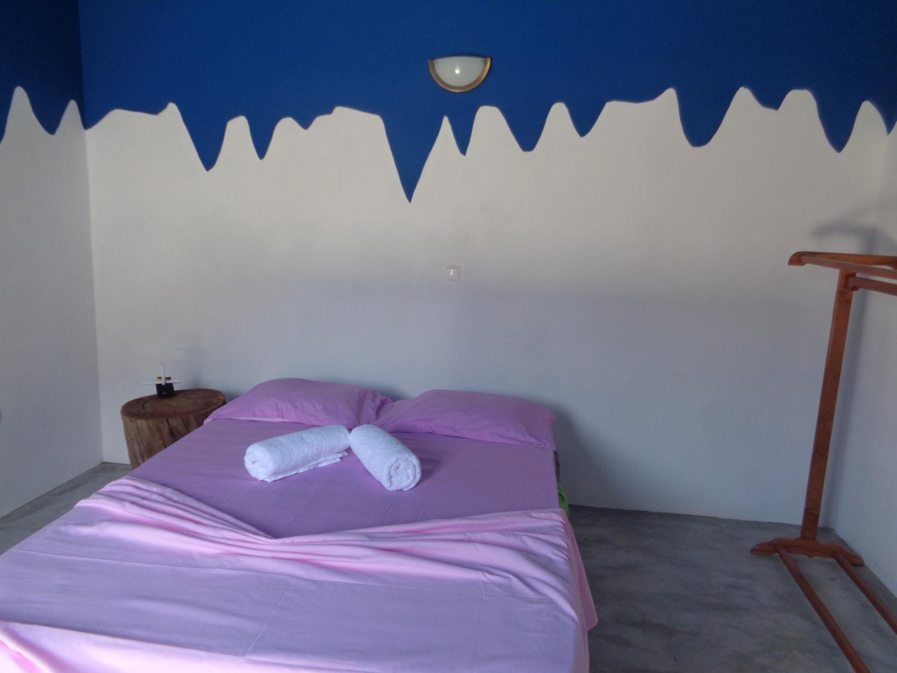 A bed with a purple bedspread and two pillows, a wooden nightstand with a small object on it, and a wooden rack against the wall.